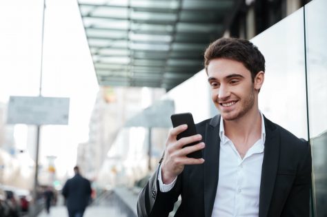 Image of smiling young businessman walking near business center. Chatting by phone. Looking aside.
